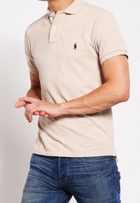 Muž v béžovém polo tričku s malým tmavým logem na hrudi a modrými džíny, stojící se spojenýma rukama před sebou.