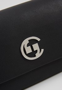 Gros plan d'une surface en cuir noir texturé avec un logo métallique circulaire argenté présentant des découpes géométriques.