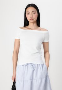 Tricou alb cu umeri descoperiți, cu mâneci scurte, textură netedă și design simplu. Asortat cu o fustă ușor albastră, cu dungi, în straturi.