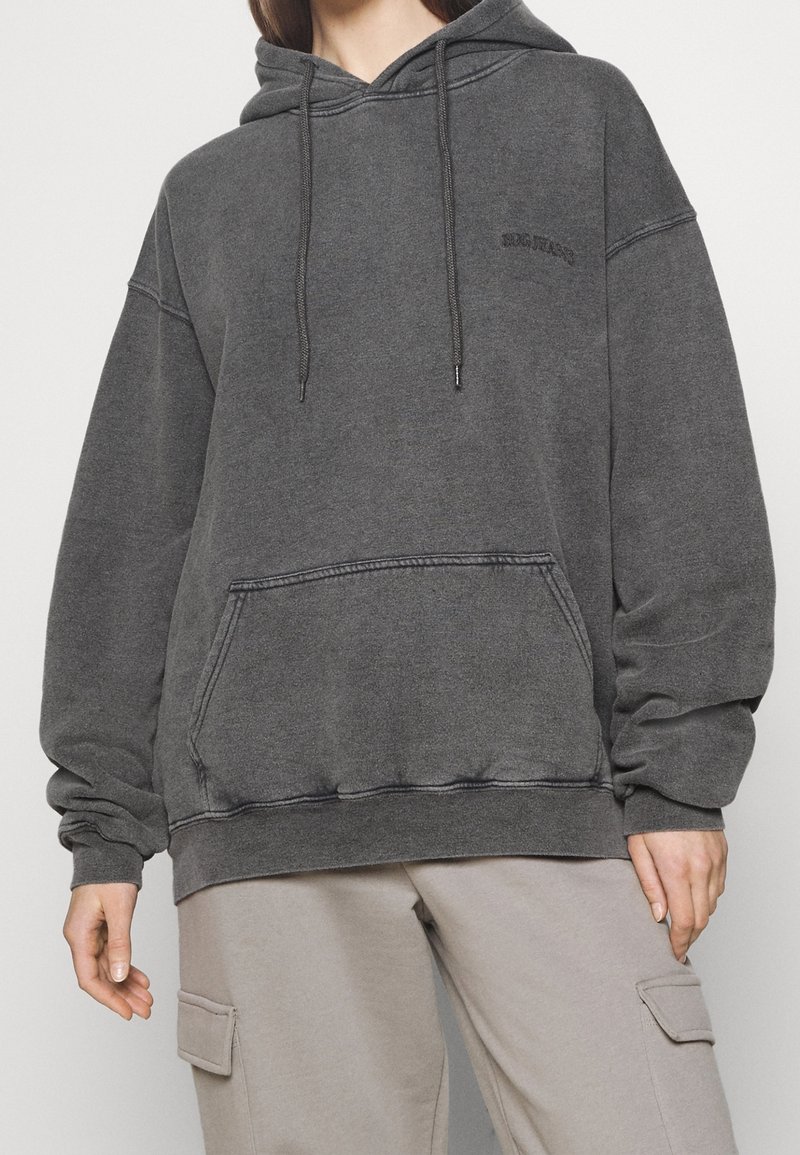 Persona che indossa un ampio hoodie nero sbiadito con tasca frontale e pantaloni cargo beige, isolata su uno sfondo chiaro uniforme.