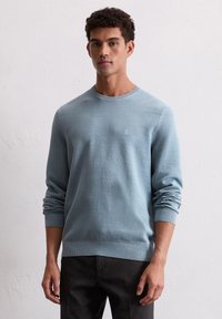 Pull en maille texturée bleu clair avec un col rond et des manches longues. Présente des poignets et un ourlet côtelés, ainsi qu'un logo discret sur la poitrine.