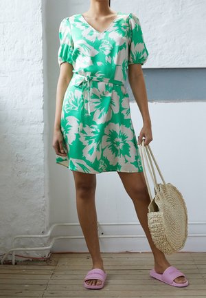 Kobieta w zielono-białej sukience w kwiaty z bufkami na rękawach, trzymająca okrągłą plecioną torebkę, stojąca na drewnianej podłodze w różowych klapkach na obcasie.