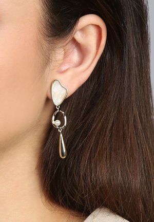 Ohr einer Frau, die einen baumelnden Ohrring mit einem unregelmäßigen weißen Stein, einer kleinen Perle in einer goldenen Schlaufe und einem goldenen Tropfenanhänger trägt.