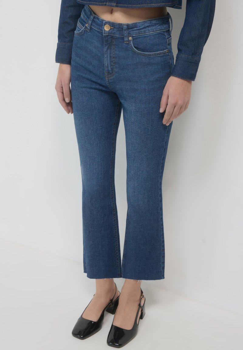 Jeans in denim a campana di colore blu scuro con vita alta, dotati di tasche frontali, chiusura con bottone e orlo grezzo alle caviglie.