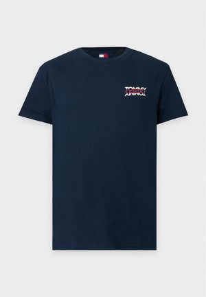 Un mannequin masculin se tient devant un fond uni, portant un T-shirt bleu marine avec logo, un jean bleu ample et des baskets blanches, une main dans la poche.