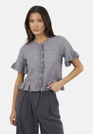 Vrouw draagt een grijs korte mouw cardigan met ruches en bijpassende grijze wijde broek tegen een effen achtergrond.