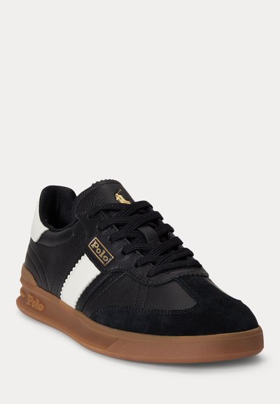 Black and white Polo sneaker with gum sole, black laces, and gold Polo logo on side and tongue, angled to show outer side.
