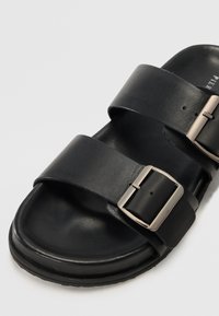 Sandal cu talpă plată din piele neagră, cu două curele late, detaliu cu cataramă metalică argintie și interior texturat pentru confort. Talpă din cauciuc pentru un plus de confort.