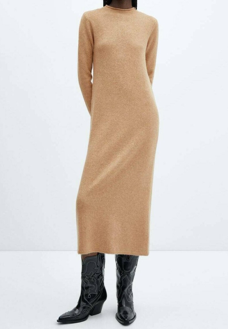 Femme portant une longue robe en maille beige associée à des bottes de cowboy en cuir noir embossé mi-mollet, debout devant un fond uni.