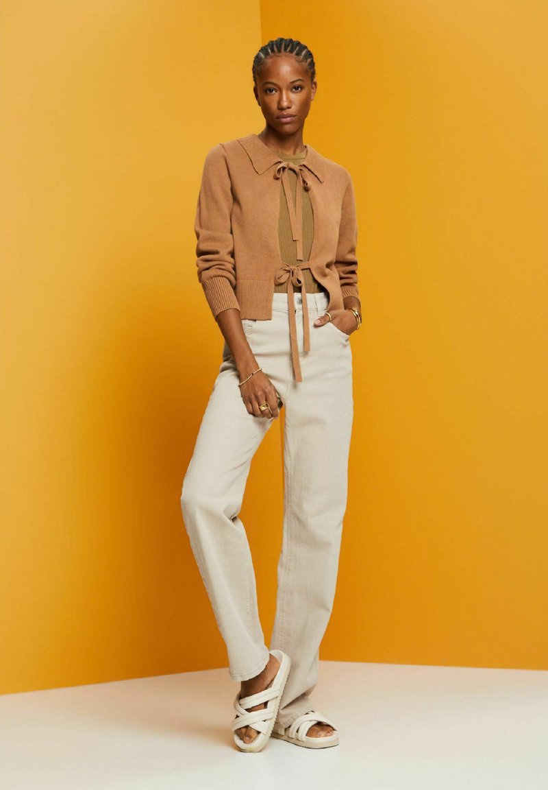 Une femme se tient contre un mur de couleur jaune moutarde, portant un cardigan marron noué sur un sweater, un pantalon beige clair, des sandales blanches, avec une main dans la poche.