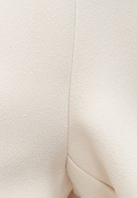 Κοντινό πλάνο σε μπεζ υφασμάτινη υφή με ορατή ραφή και γωνιακή δίπλωση που δημιουργεί διακριτική σκιά.