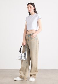 Jonge vrouw staat zelfverzekerd in een cropped lila top en baggy beige jeans, met een glanzende zilveren Calvin Klein handtas in haar hand. Effen achtergrond.