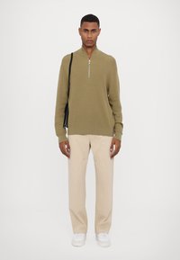 Olive green half-zip sweater with ribbed texture, paired with beige straight-leg pants. White sneakers complete the outfit. Simple black bag worn.