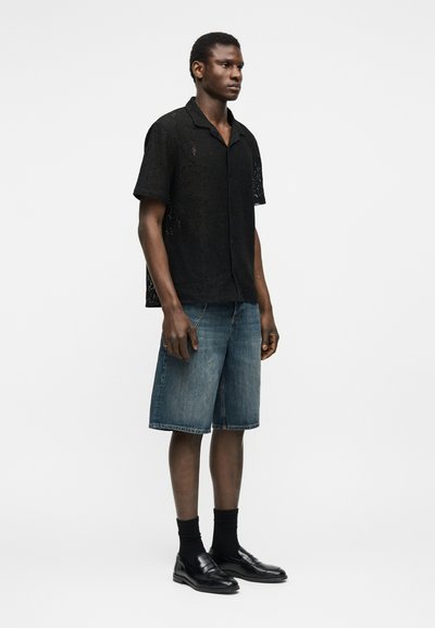 Άνδρας που στέκεται φορώντας μαύρο δαντελένιο πουκάμισο με κοντά μανίκια, τζιν σορτς μήκους μέχρι το γόνατο, μαύρες κάλτσες και μαύρα δερμάτινα μοκασίνια σε λευκό φόντο.