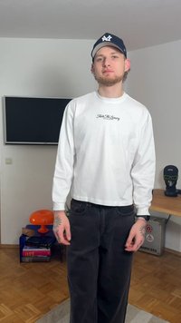 Vit långärmad skjorta med texten "Taste The Luxury", kombinerad med svarta jeans med vida ben. Personen bär en mörk keps och har synliga tatueringar.