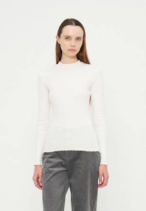 Jeune femme aux cheveux bruns et lisses portant un haut blanc à manches longues et un pantalon gris, debout devant un fond clair uni.