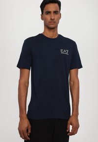 Námořnická modrá tričko s krátkým rukávem vyrobené z měkké bavlny, s kulatým výstřihem a bílým logem EA7 na levé části hrudi. Hladká textura.