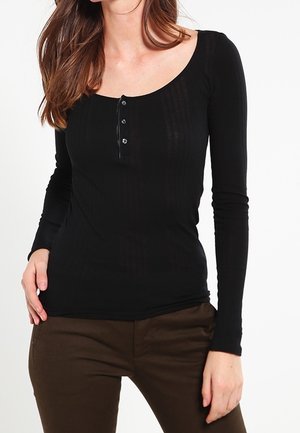 Kobieta ubrana w dopasowaną czarną bluzkę z długim rękawem z trzema guzikami z przodu oraz brązowe spodnie, stojąca na białym tle.