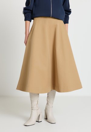 Frau trägt beigen A-Linien-Midirock kombiniert mit weißen kniehohen Stiefeln mit Absatz und navyblauer Jacke mit Reißverschluss.