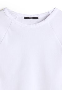 Camiseta de manga larga blanca con cuello redondo y mangas raglán. La etiqueta de la marca es visible cerca del cuello. Textura suave.
