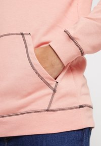 Felpa rosa con tasca a marsupio, cuciture nere in evidenza e una texture morbida. I polsini e l'orlo presentano dettagli di cucitura coordinati.