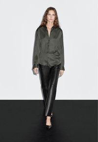 Donkergroene satijnen blouse met knopen en kraag, gecombineerd met op maat gemaakte zwarte leren broek en elegante zwarte hakken, gefotografeerd tegen een eenvoudige achtergrond.