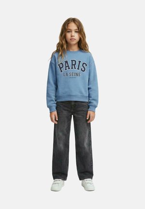 Jong meisje met lang golvend haar, een blauw "PARIS LA SEINE" sweatshirt, donkere wijd uitlopende jeans en witte sneakers, staand en recht vooruit kijkend.
