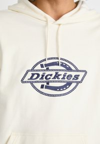 Kremfarget hettegenser med en stor, marineblå sirkulær grafikk som har ordet "Dickies" tydelig brodert i sentrum. Myk tekstur.