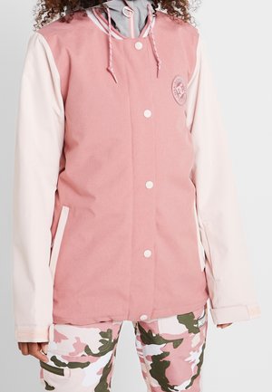 Osoba ubrana w różowo-kremową kurtkę z kapturem i zapięciem na guziki oraz różowo-zielono-białe spodnie moro, stojąca na jednolitym tle.