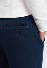 Mörkblå sweatpants med ribbad textur, sidoficka med röda accenter och en märkesetikett på baksidan. Komfortabelt tyg.