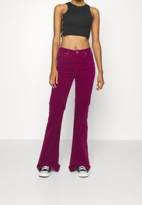 Valkoiset, pörröiset samettihousut kirkkaassa fuksiassa, joissa on korkea vyötärö ja teksturoitu kangas, yhdistetty mustaan ribbattuun crop-toppiin ja tennareihin.