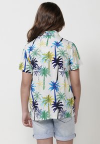 Camisa de manga corta con un patrón de palmeras en verde, azul y amarillo sobre un fondo blanco; tejido ligero con un corte relajado.