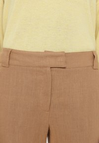 Gros plan d'un pantalon en lin beige avec une ceinture recouverte de tissu et une fermeture avant cachée, porté avec un pull jaune pâle.