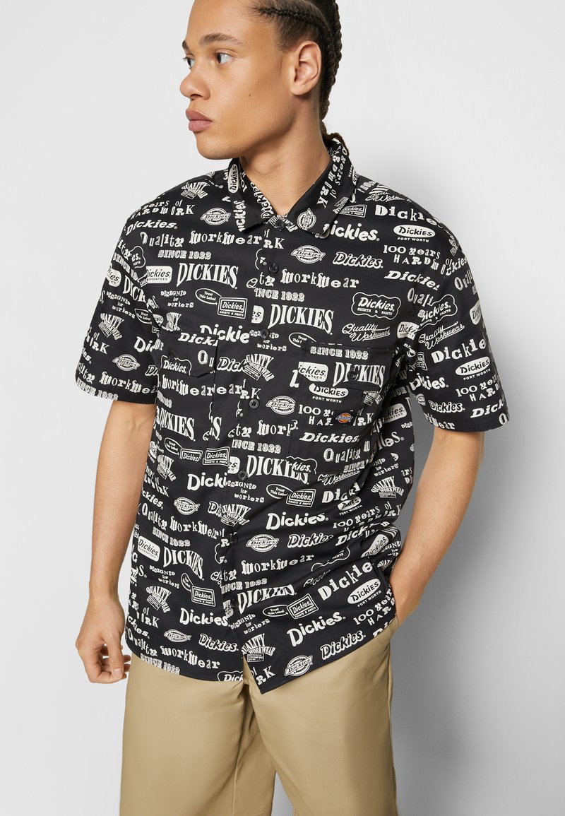 Short-sleeve black shirt featuring various white logos and text patterns. Collar and button closure, made of lightweight fabric. Khaki pants worn.