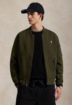 Jeune homme portant une casquette noire, une veste zippée vert olive avec un logo blanc, un pull noir et un pantalon noir, debout devant un fond uni.