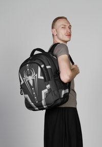 Sac à dos noir avec un design d'araignée en gris, en tissu texturé, sangles rembourrées et plusieurs compartiments pour le rangement.