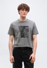 Νέος άνδρας με σγουρά μαλλιά που φοράει γκρι μπλούζα με μαύρα σχέδια δακτυλίων δέντρου και τη λέξη «BOSS», συνδυασμένη με μαύρο παντελόνι.