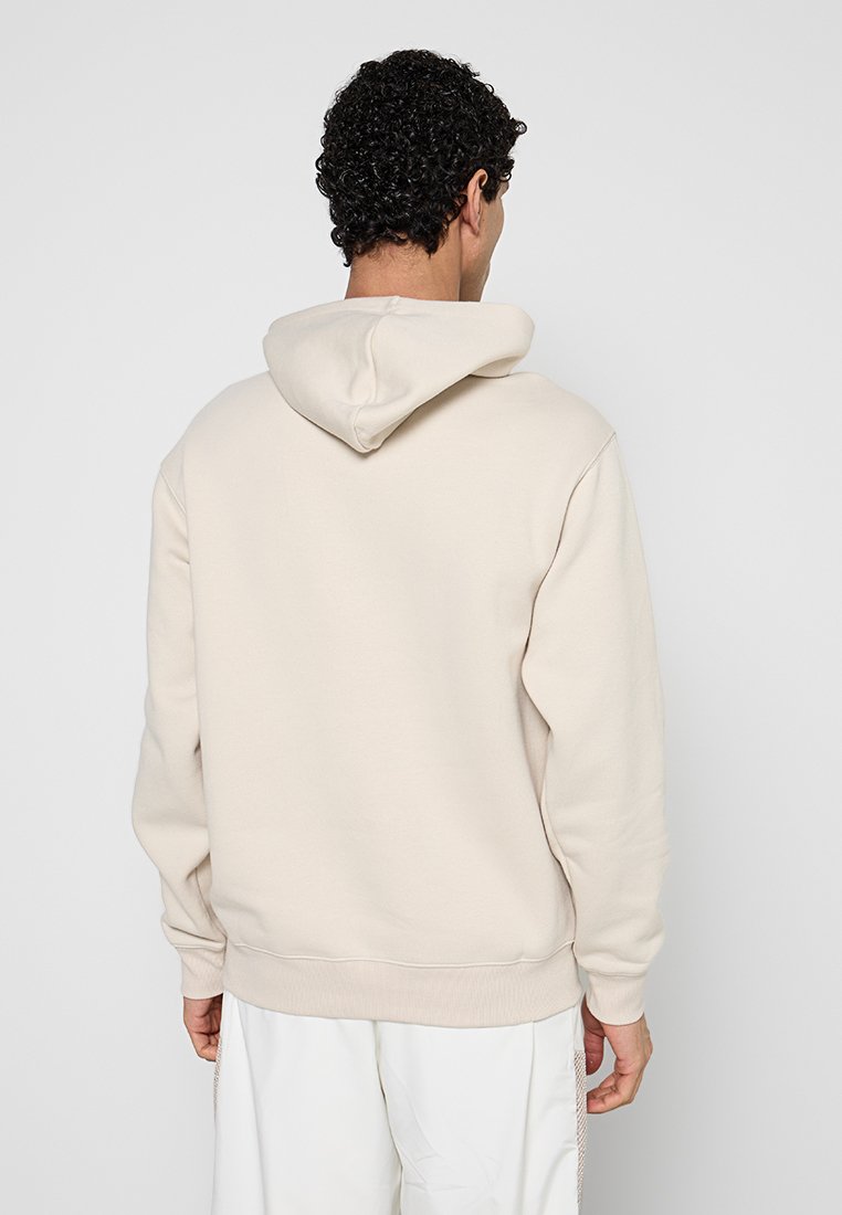 Mann mit lockigem Haar, der einen hellbeigen Hoodie und weiße Hosen trägt und mit dem Rücken zum Betrachter vor einem schlichten grauen Hintergrund steht.