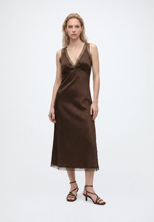 Mulher vestindo um vestido midi castanho de cetim sem mangas com acabamento em renda e sandálias castanhas de salto com tiras sobre fundo branco.