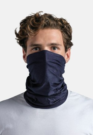 Young man with curly hair wearing a dark blue neck gaiter covering nose and mouth, dressed in a white shirt, neutral background.