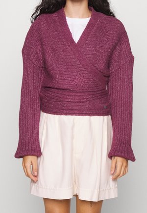 Femme portant un pull cache-cœur violet côtelé à manches longues sur un haut blanc et un short beige clair, debout devant un fond uni.