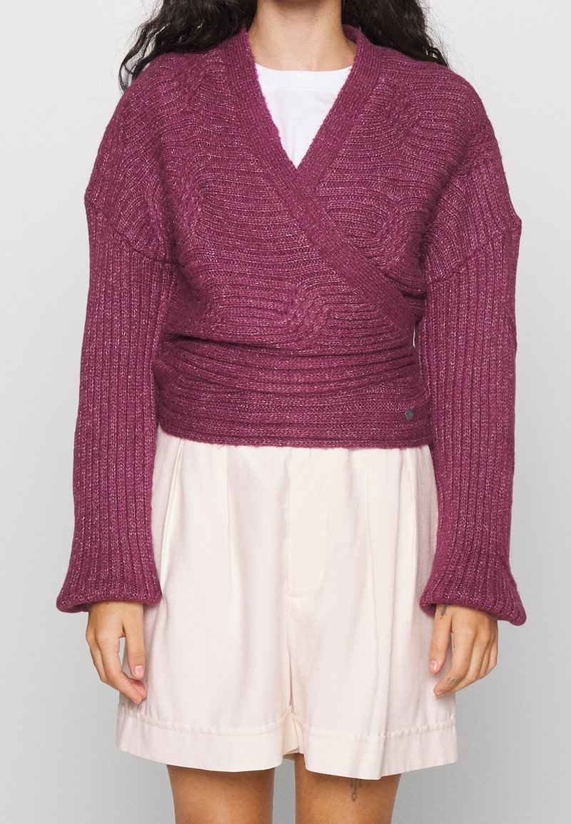 Femme portant un pull cache-cœur violet côtelé à manches longues sur un haut blanc et un short beige clair, debout devant un fond uni.