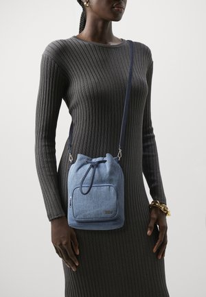 Personne portant une robe à manches longues côtelée avec un bracelet en or et portant un petit sac bandoulière bleu à cordon avec une poche zippée à l'avant.