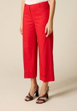 Mujer vistiendo pantalones anchos y cortos de color rojo brillante, combinados con sandalias de tacón alto de color marrón oscuro, de pie frente a un fondo neutro.