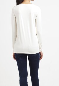 Långärmad, krämfärgad topp med rund halsringning. Mjuk textur och figurnära design, ihop med mörkblå tajta jeans.