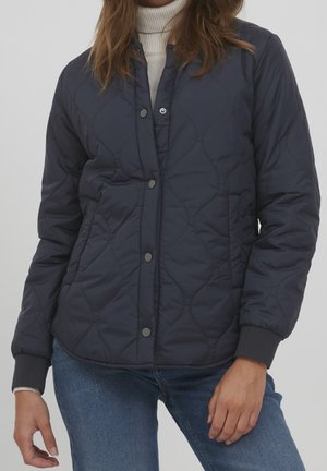 Person klädd i en marinblå quiltad jacka med tryckknappar över en vit polotröja och blåa jeans, stående mot en enfärgad bakgrund.