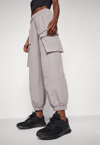 Persona che indossa pantaloni jogger cargo grigio chiaro con polsini elasticizzati e scarpe da ginnastica nere, in piedi su uno sfondo semplice.