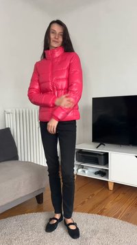 Jasnoróżowa puchowa kurtka z wysokim kołnierzem i gładką fakturą, w połączeniu z czarnymi przylegającymi spodniami i czarnymi płaskimi butami.