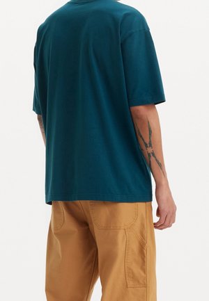 Man som bär en överdimensionerad t-shirt i teal och beige byxor, visar en svart klo-liknande tatuering på sin vänstra underarm.