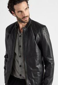 Zwarte leren jas met gestikte schouderaccenten, staande kraag, ritssluiting en platte naden. Gedragen over een gestreept overhemd met knopen.